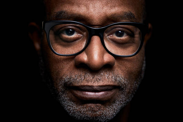 Close-up portrait of an adult man of African descent wearing glasses, with a neutral expression, against a dark background.