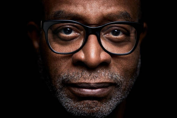 Close-up portrait of an adult man of African descent wearing glasses, with a neutral expression, against a dark background.