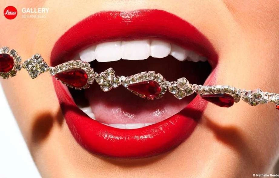 Close-up studio shot of a woman's red lipstick-covered mouth with open teeth, and holding a jeweled bracelet between her teeth.