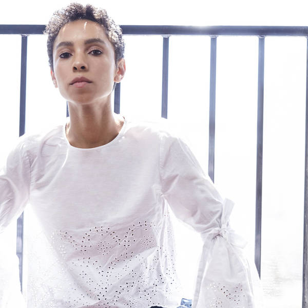 Elegant woman with short curly hair poses indoors wearing a white blouse with bell sleeves and decorative perforations with sunlight.