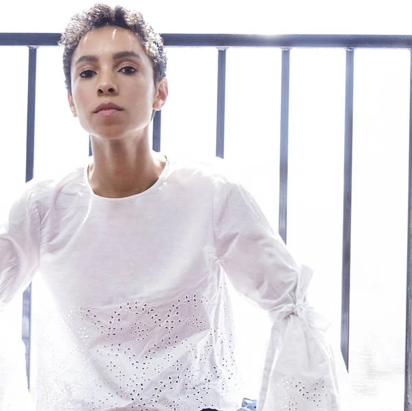 Elegant woman with short curly hair poses indoors wearing a white blouse with bell sleeves and decorative perforations with sunlight.