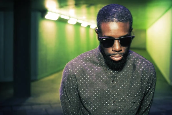 Portrait of a Black man with a beard and sunglasses in a tunnel with green lighting, showcasing a blend of urban and personal style.