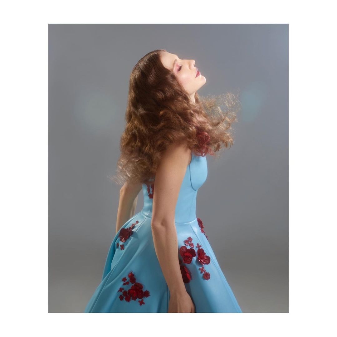 Side profile of a woman with long, wavy auburn hair, fair skin, closed eyes, and light pink lipstick wearing a blue dress with red floral accents.