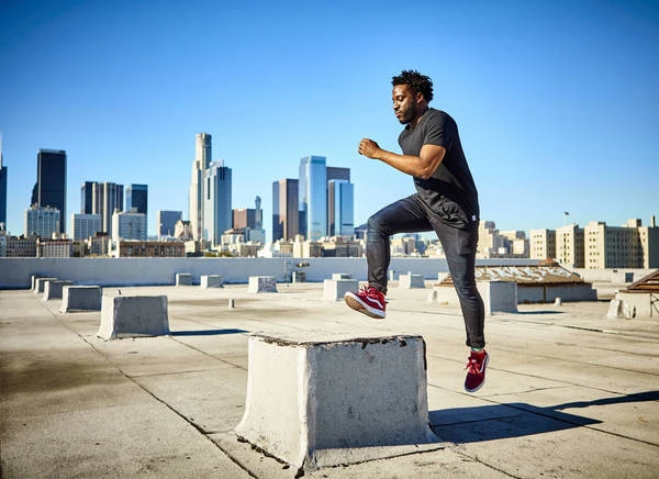Athletic Black man steps onto a concrete block on a rooftop with a cityscape backdrop, showcasing fitness and urban athleticism against a blue sky.