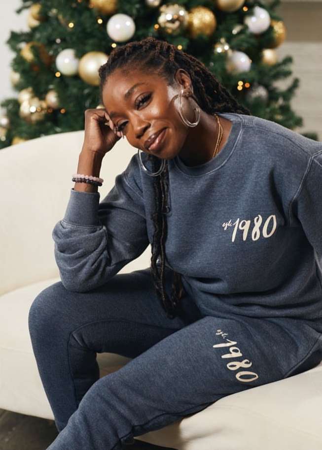 Adult woman in grey sweatshirt and sweatpants poses on a white couch with a Christmas tree in the background, medium shot.