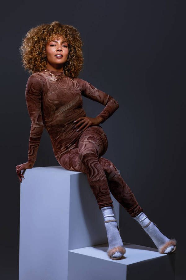 A woman with curly hair poses while sitting on white blocks in a long-sleeved bodysuit with a swirled brown pattern in a studio setting.