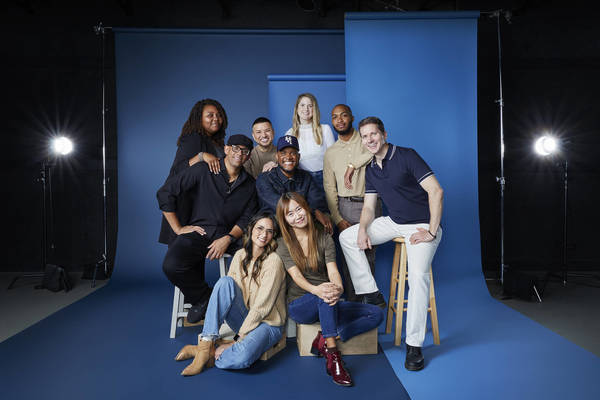 Group photo of nine diverse adults in a studio setting, seated and standing against a blue backdrop with studio lighting, smiling.