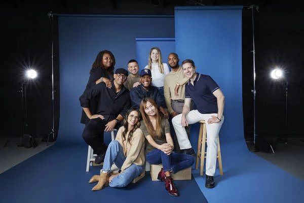 Group photo of nine diverse adults in a studio setting, seated and standing against a blue backdrop with studio lighting, smiling.