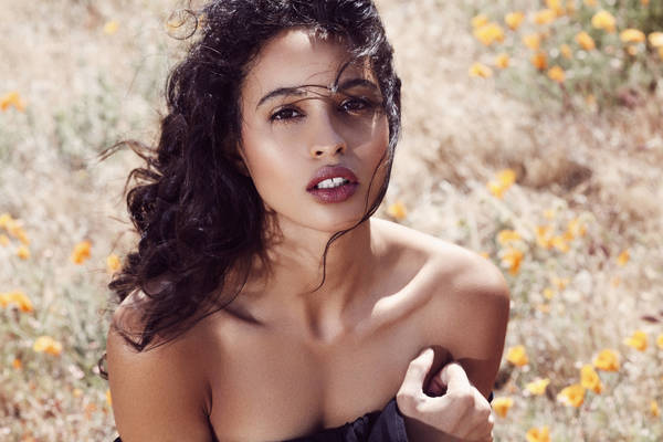 Portrait of a young woman with dark hair, posing outdoors among orange wildflowers in a field, bathed in soft natural light.