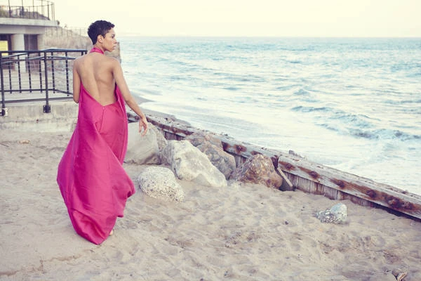 Woman in a long magenta backless dress walking on a sandy beach near the ocean by Nathalie Gordon