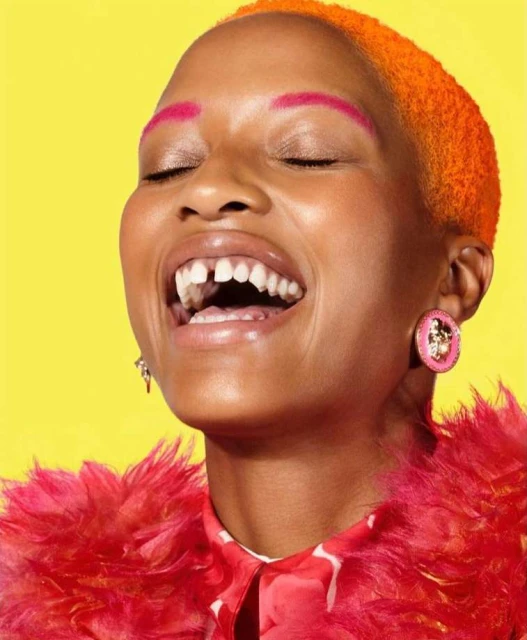 Close-up portrait of a smiling African-American woman with orange hair, pink eyebrows, and pink and red feathered clothing against a yellow background.