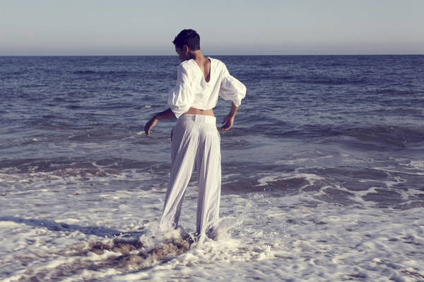Woman stands in the ocean surf wearing a white blouse and striped pants against a background of sea and sky.