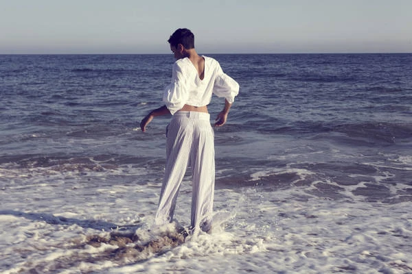 Woman stands in the ocean surf wearing a white blouse and striped pants against a background of sea and sky.