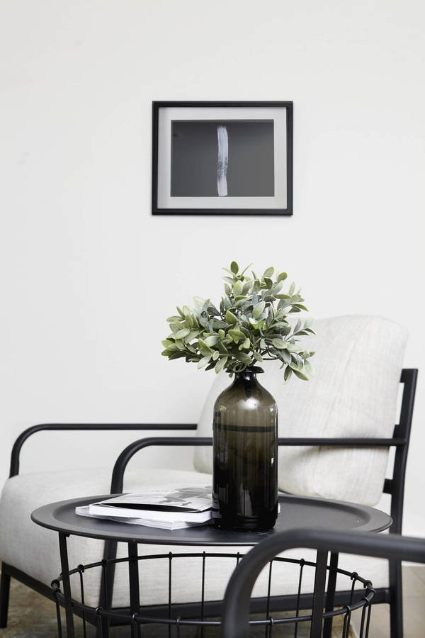 Interior design featuring a leafy plant in a bottle vase on a black metal table next to a modern armchair and framed art print.