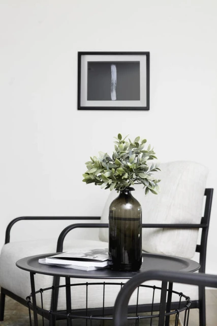 Interior design featuring a leafy plant in a bottle vase on a black metal table next to a modern armchair and framed art print.
