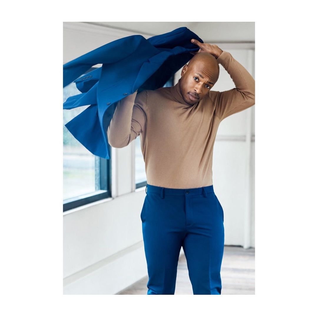 Adult male in a tan shirt and blue pants putting on a blue jacket in an indoor setting with natural lighting from nearby windows.