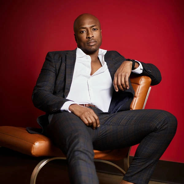 Portrait of an adult male wearing a suit. He's sitting in a leather chair against a solid red background in a photo studio.