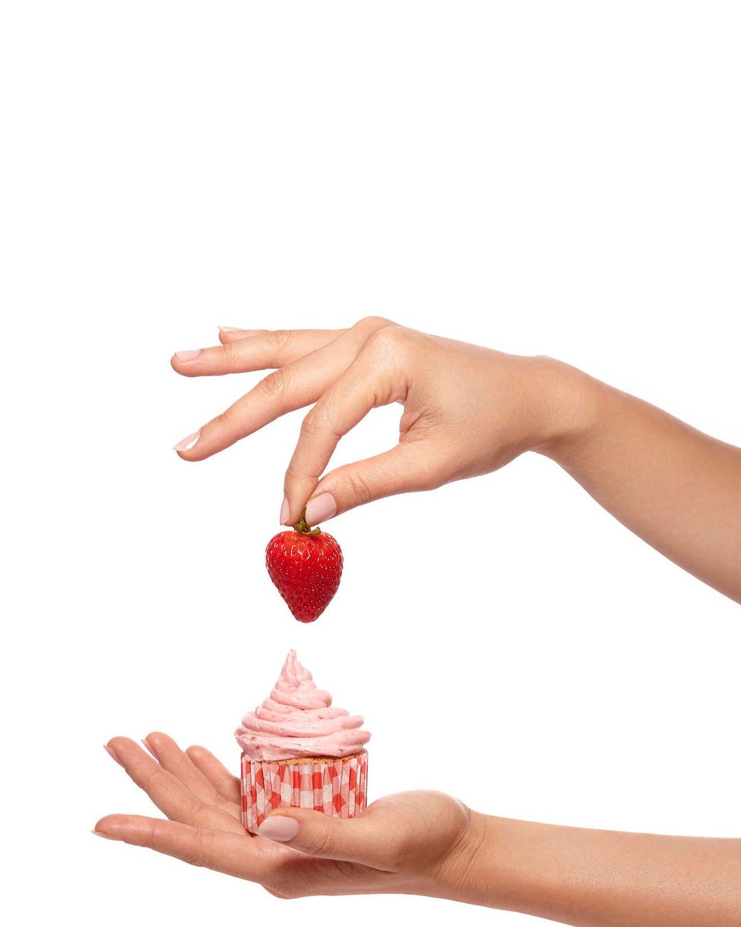Close-up shot of a person's hands holding a cupcake with pink frosting and placing a strawberry on top, studio shot against white.