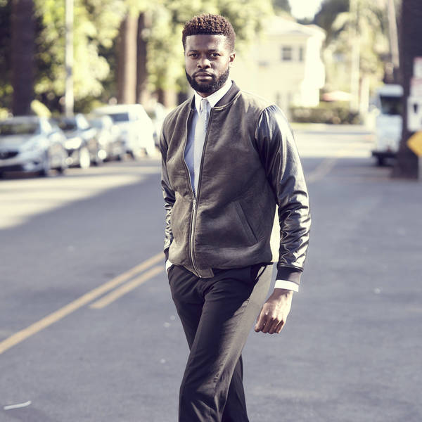 A stylish adult male with dark hair walks down an outdoor city street, wearing a leather bomber jacket, white shirt, and dark slacks.