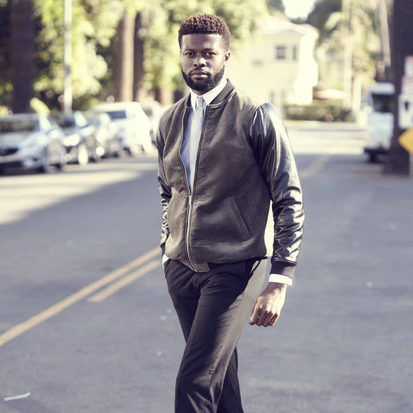 A stylish adult male with dark hair walks down an outdoor city street, wearing a leather bomber jacket, white shirt, and dark slacks.