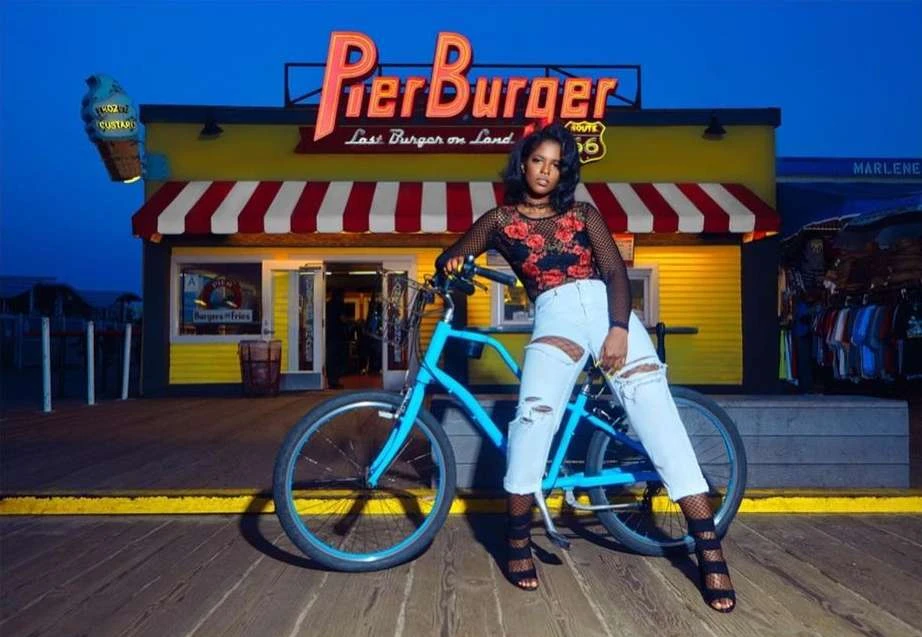 Woman poses with a bike in front of the PierBurger restaurant at dusk, wearing a mesh top, jeans, and heels, with the restaurant signage visible.