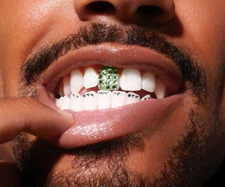 Close-up of a man's mouth showing teeth with green and silver dental grills, finger slightly touching his lip.