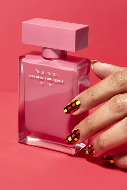 Close-up of a Fleur Musc Narciso Rodriguez perfume bottle on a red background with a woman's hand displaying patterned nails.