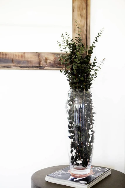 Eucalyptus branches in a tall clear glass vase on a grey side table with a magazine, against a white wall with wood-frame art.