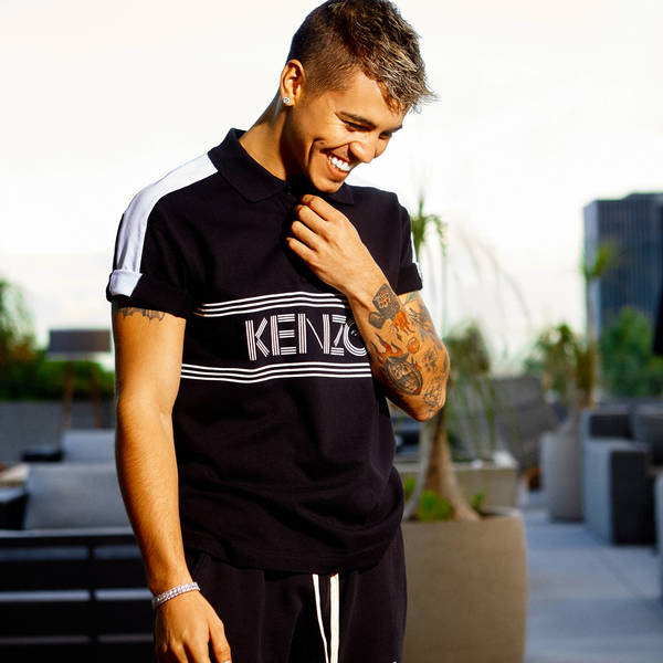 Young man smiles, wearing a black Kenzo polo shirt with white stripes and sleeves, on a rooftop terrace with a city skyline view.