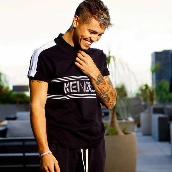 Young man smiles, wearing a black Kenzo polo shirt with white stripes and sleeves, on a rooftop terrace with a city skyline view.
