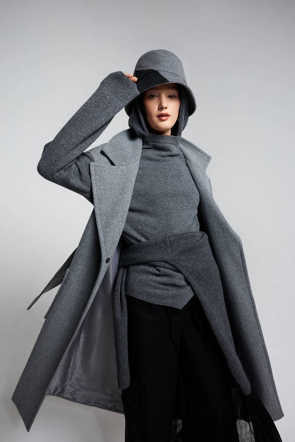 Stylish young woman in grey coat, matching turtleneck, and bucket hat posing against a plain grey background in a fashion shot.