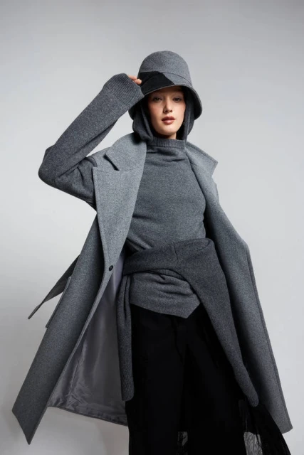 Stylish young woman in grey coat, matching turtleneck, and bucket hat posing against a plain grey background in a fashion shot.
