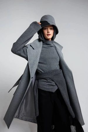 Stylish young woman in grey coat, matching turtleneck, and bucket hat posing against a plain grey background in a fashion shot.
