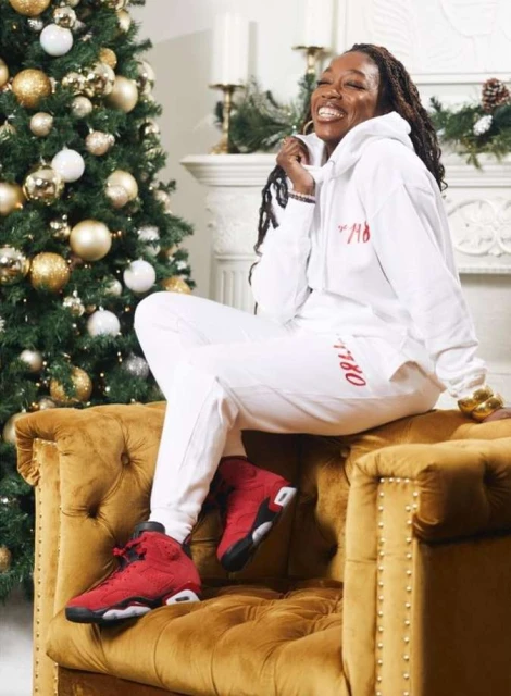 Stylish woman in athleisure wear poses on a velvet chair with a Christmas tree, showcasing red sneakers and gold accessories.