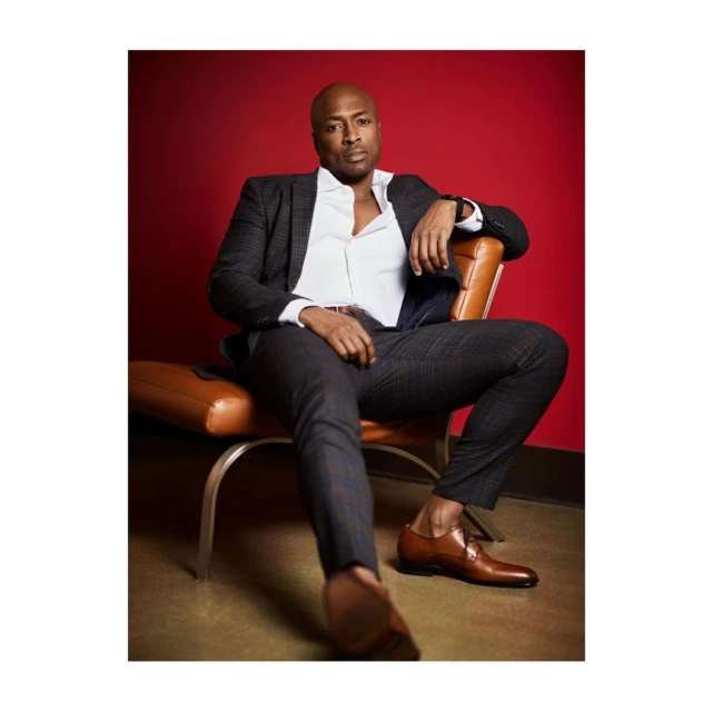Stylish adult male seated in a chair wearing a suit, open-collar shirt, and dress shoes against a red backdrop.