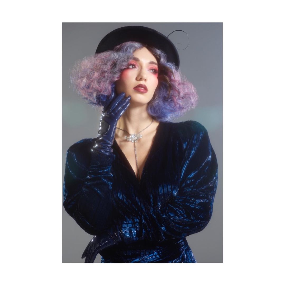 Elegant woman in blue velvet dress, gloves, and hat. She has striking makeup and multicolored hair, posing against a grey backdrop.