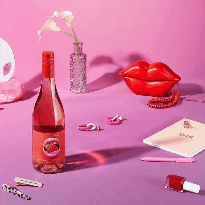 Still life with a bottle of strawberry juice, a lips-shaped telephone, and various pink accessories on a pink and purple surface.