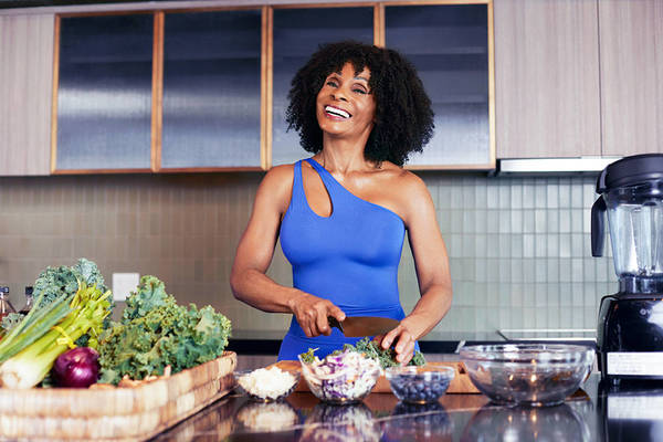 Adult female in blue activewear smiles while chopping greens in a kitchen with stainless steel appliances and a blender.