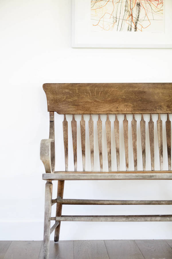 A wooden bench sits against a white wall with an abstract framed picture above it and a dark hardwood floor below it.