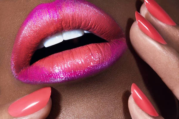 Close-up of a woman's lips with vibrant fuchsia to coral gradient lipstick, complemented by long, coral-painted nails.