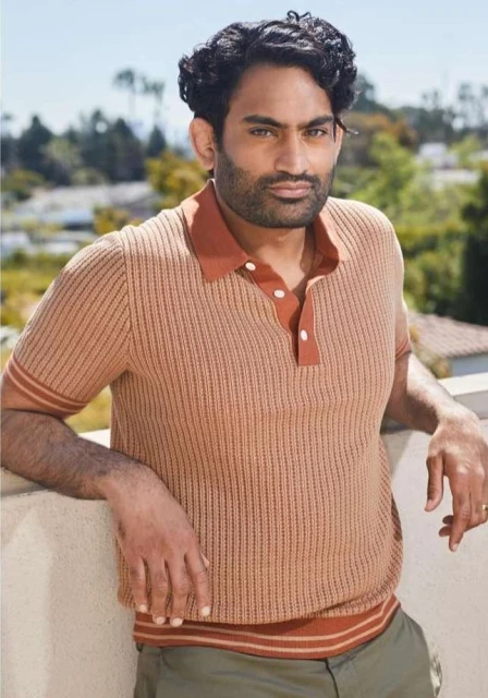 Portrait of a man with dark hair and beard, wearing a striped polo shirt, leaning against a white wall, with a green outdoor background.