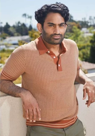 Portrait of a man with dark hair and beard, wearing a striped polo shirt, leaning against a white wall, with a green outdoor background.