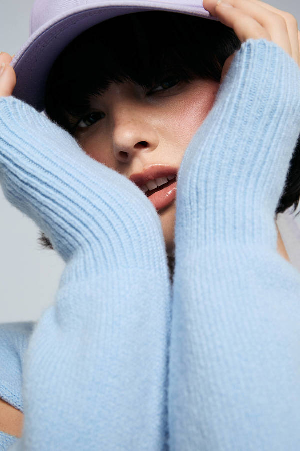 Close-up of a young adult woman with fair skin wearing a light blue sweater and lavender baseball cap, holding her hands to her face.