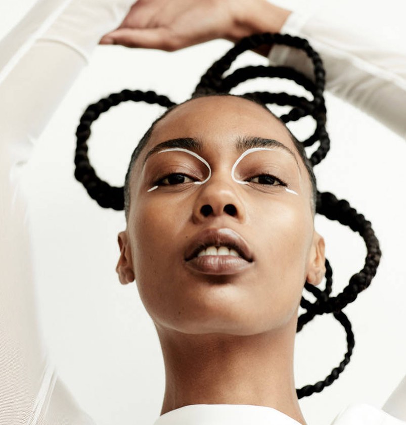 Close-up portrait of a woman with braided hair, striking white eyeliner, and hands posed above her head against a white background.