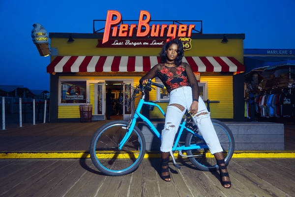 A woman in ripped jeans and a floral top leans on a teal bicycle in front of Pier Burger in an outdoor, nighttime setting.
