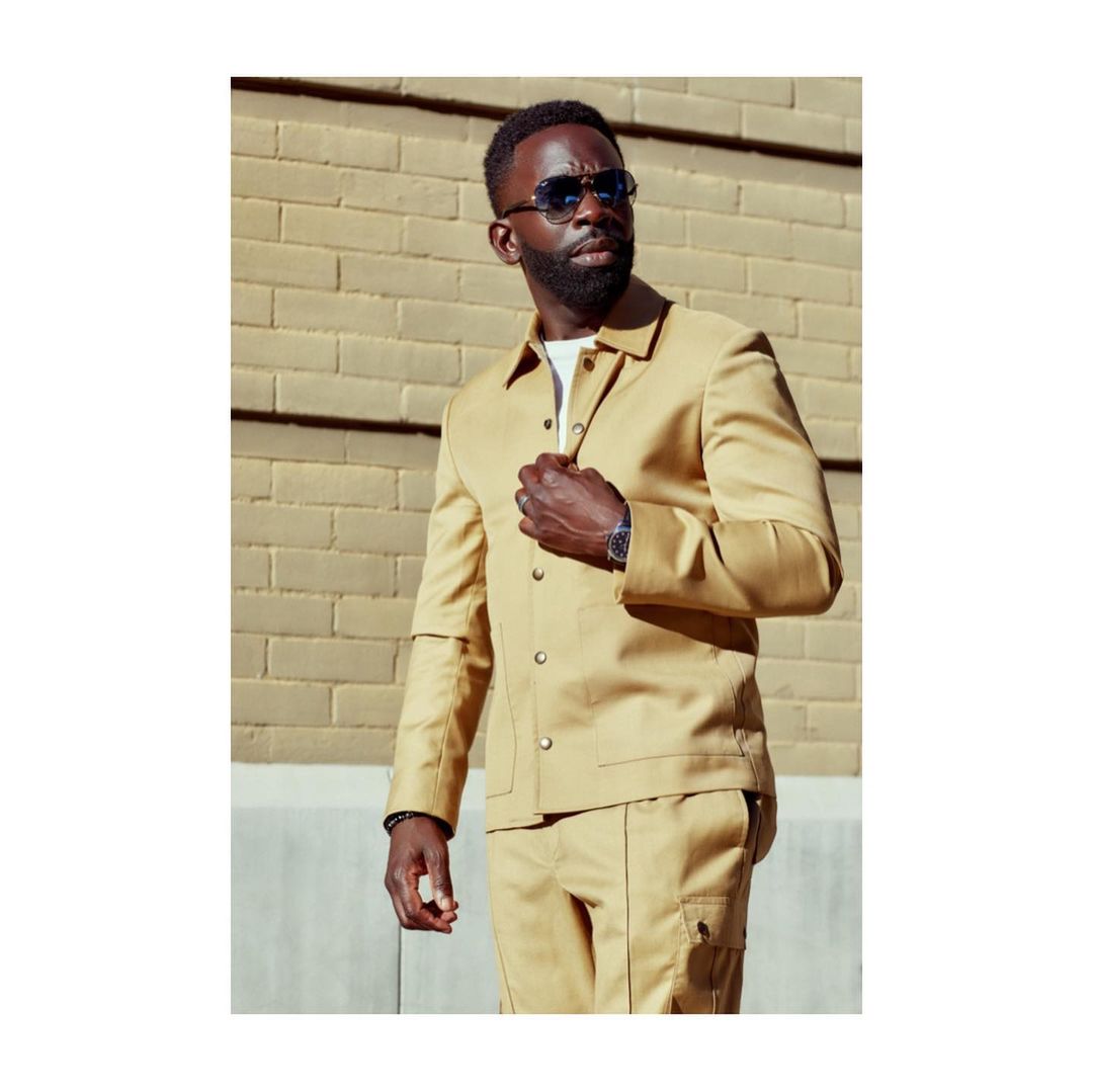 An African-American man in a tan jacket and pants poses against a brick wall, showcasing fashion style with sunglasses and a watch.