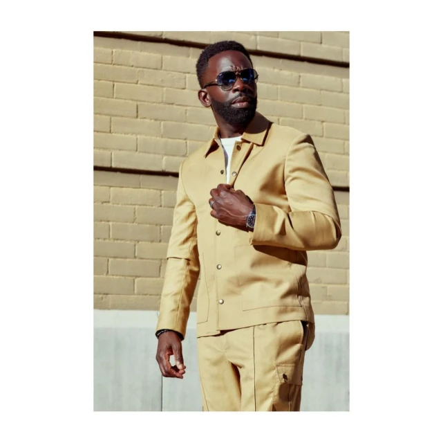 An African-American man in a tan jacket and pants poses against a brick wall, showcasing fashion style with sunglasses and a watch.