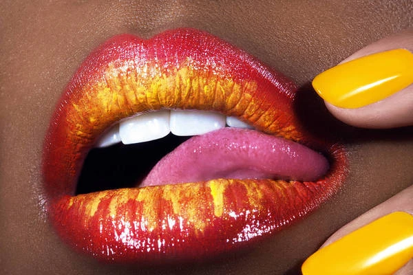 Close-up of a person's lips with vibrant red and gold lipstick, tongue slightly out, and manicured yellow fingernails visible.