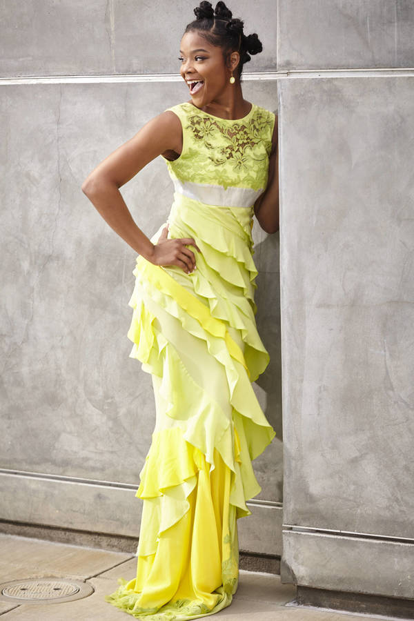 Elegant adult woman poses in a vibrant yellow gown against a textured grey wall, smiling broadly with joy. Her hair is styled in playful knots.