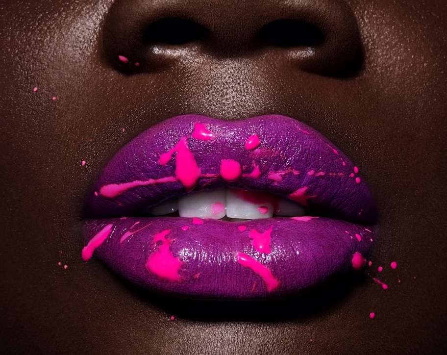 Close-up of an adult female's face showcasing vibrant purple lipstick with pink paint splatters for a bold beauty statement.